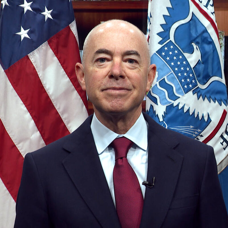 This image is of DHS Secretary Alejandro Mayorkas standing in front of a book case and two flags.