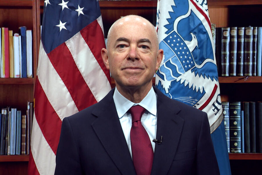 This image is of DHS Secretary Alejandro Mayorkas standing in front of a book case and two flags.