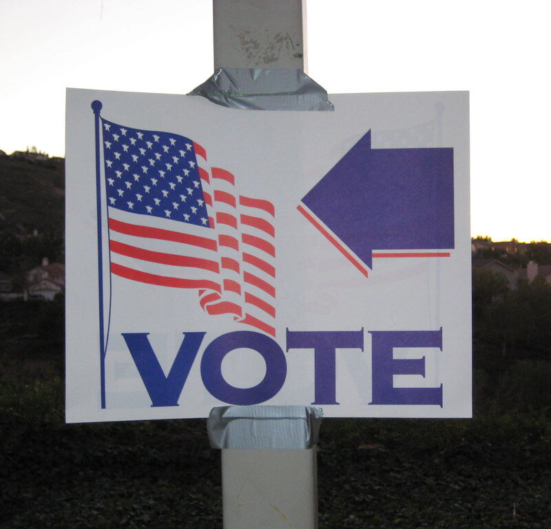 This image is of a sign that reads, "Vote" with an arrow pointing to an American Flag