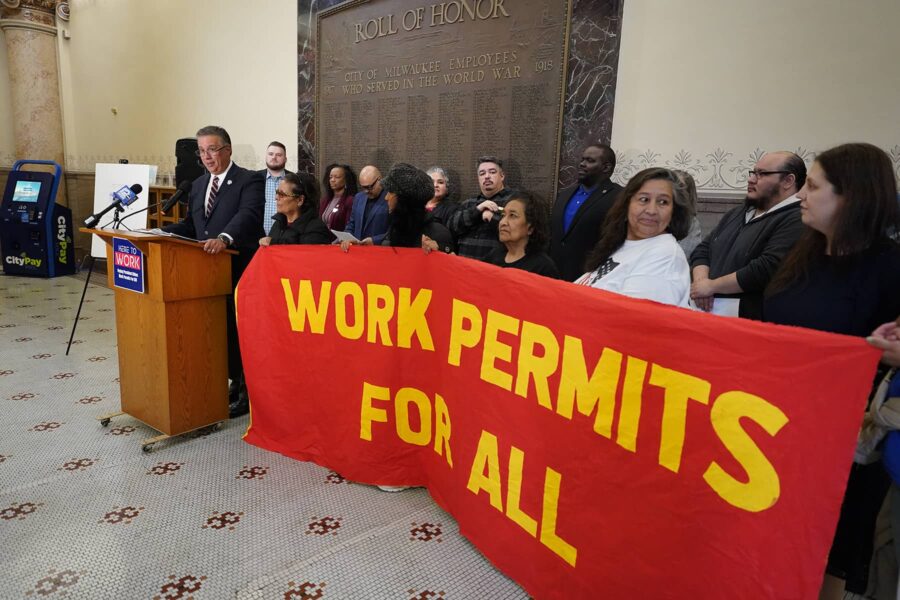 This image is of people holding a red sign that reads, "Work Permits For All" in yellow lettering.